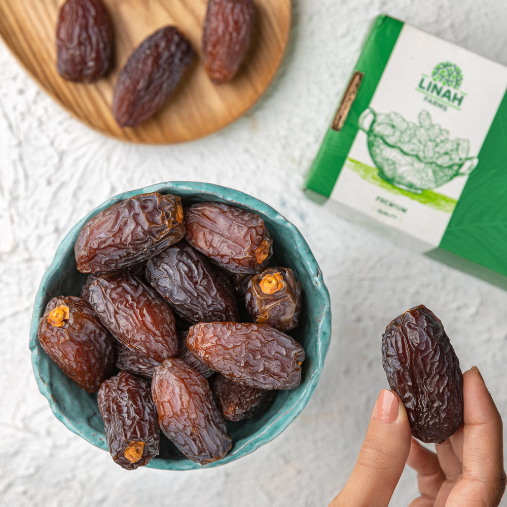 Date palm fruits in a bowl with a hand holding a date, Linah Farms packaging in the background.