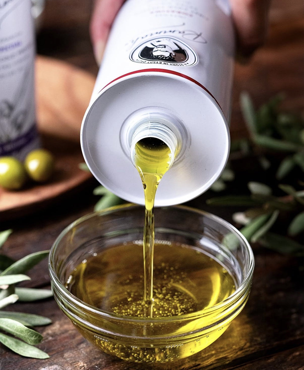 Olive oil being poured from a tin into a glass bowl with olive leaves around.